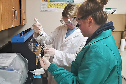 Students wearing lab coats and goggles talk in an engineering lab