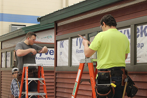 Two students on ladders work on siding and trim on the outside of a shed