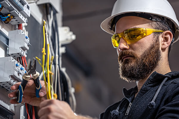 An electrician wearing protective equipment works on an electrical panel in an industrial plant