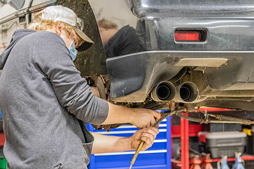 A student works on the exhaust system of a car on a lift. 