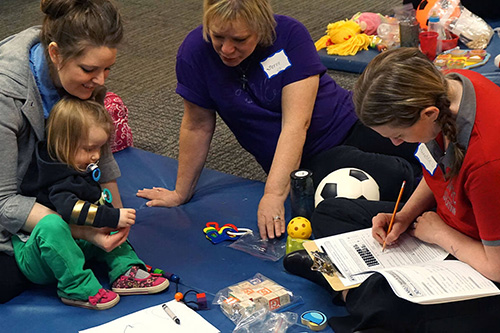 A parent, educator and an early childhood student sit with a toddler on a blanket with toys