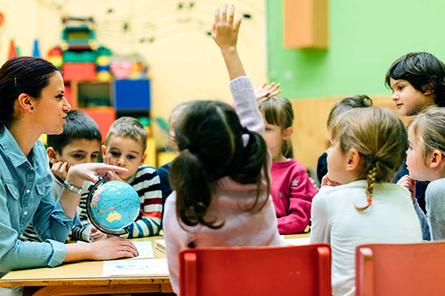 A teacher in a classroom with children raising their hands