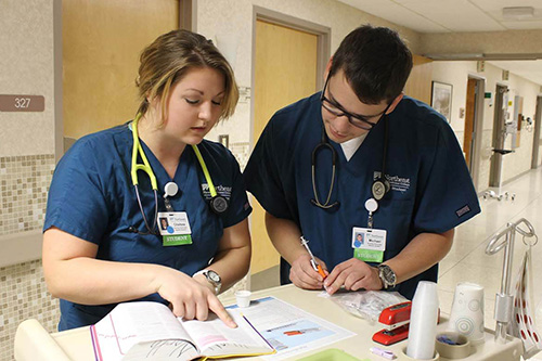 Two nurses in scrubs consult a medication handbook while distributing medications