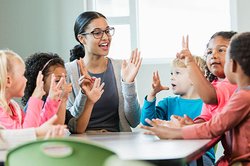 A teacher sits at a table with children raising their hands
