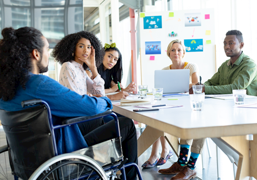 Office meeting with five people, including a wheelchair user and laptop user, around a table with planning materials