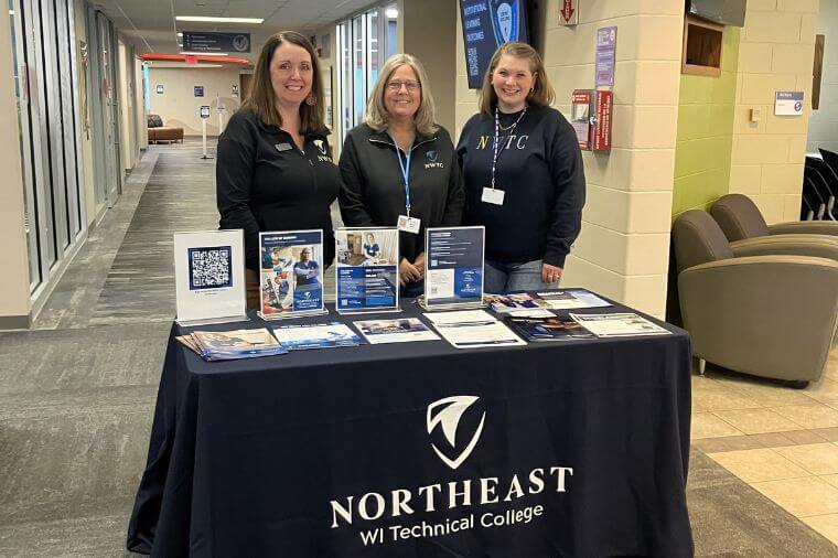 Three women standing at table with a dark blue table cloth with papers branded for Northeast Wisconsin Technical College. 