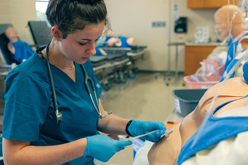 A nursing student in scrubs draws blood on a patient simulator