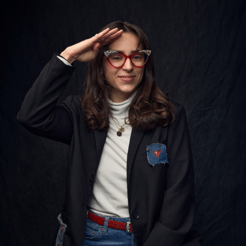A person with long hair and glasses saluting.