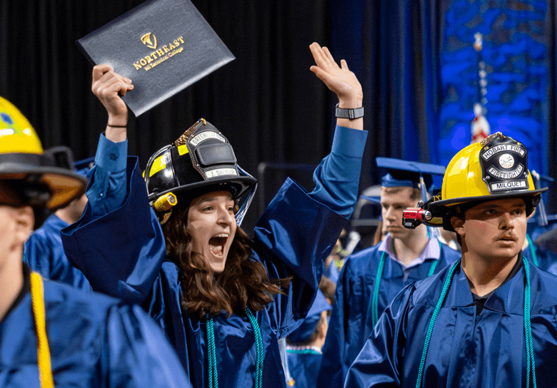Graduates in blue gowns and firefighter helmets celebrate, with one raising a diploma overhead.