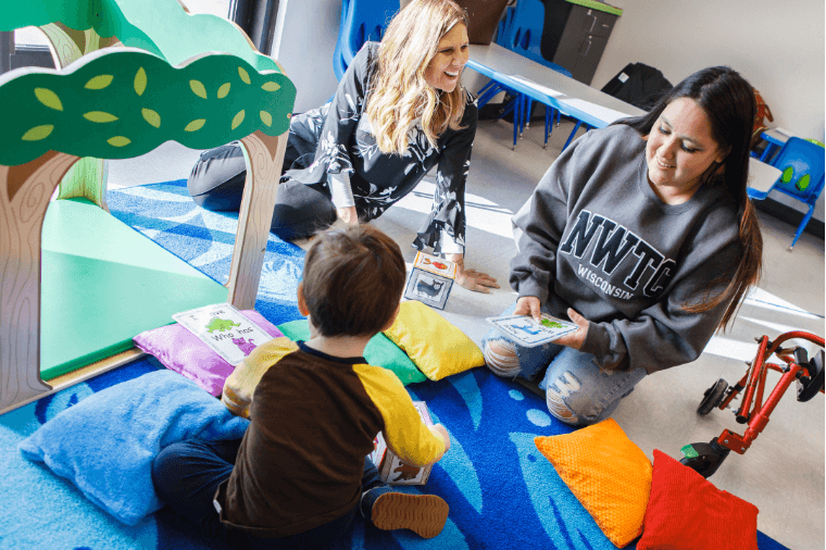 Two adults and a child interacting on a colorful rug with books and toys in a playful, educational setting.