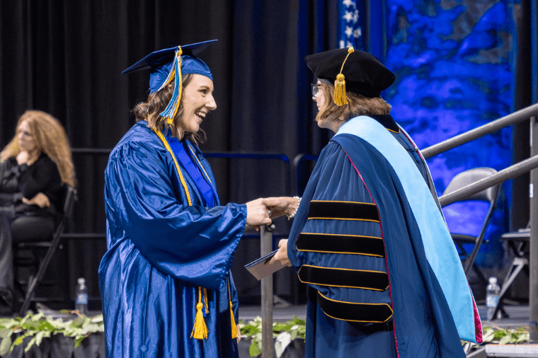 Two people in caps and gowns shaking hands and smiling on stage.