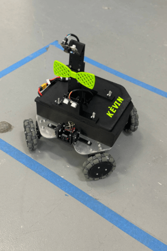 A small black robot named Kèvin is positioned on a gray surface with blue tape. The robot features a bright green bow tie attached to the front.