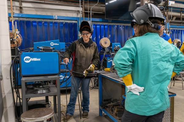 Sarah Fischer, wearing safety glasses and gear, stands in welding lab with small group of students