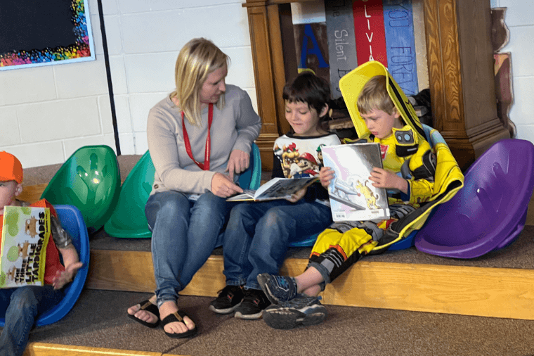 An adult and two children are sitting and reading books together in a colorful classroom setting.
