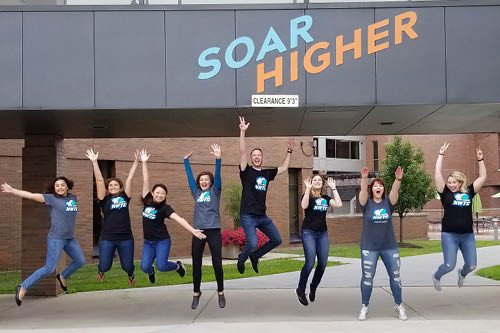 Recruitment team jumps below a welcome sign wearing NWTC tee shirts
