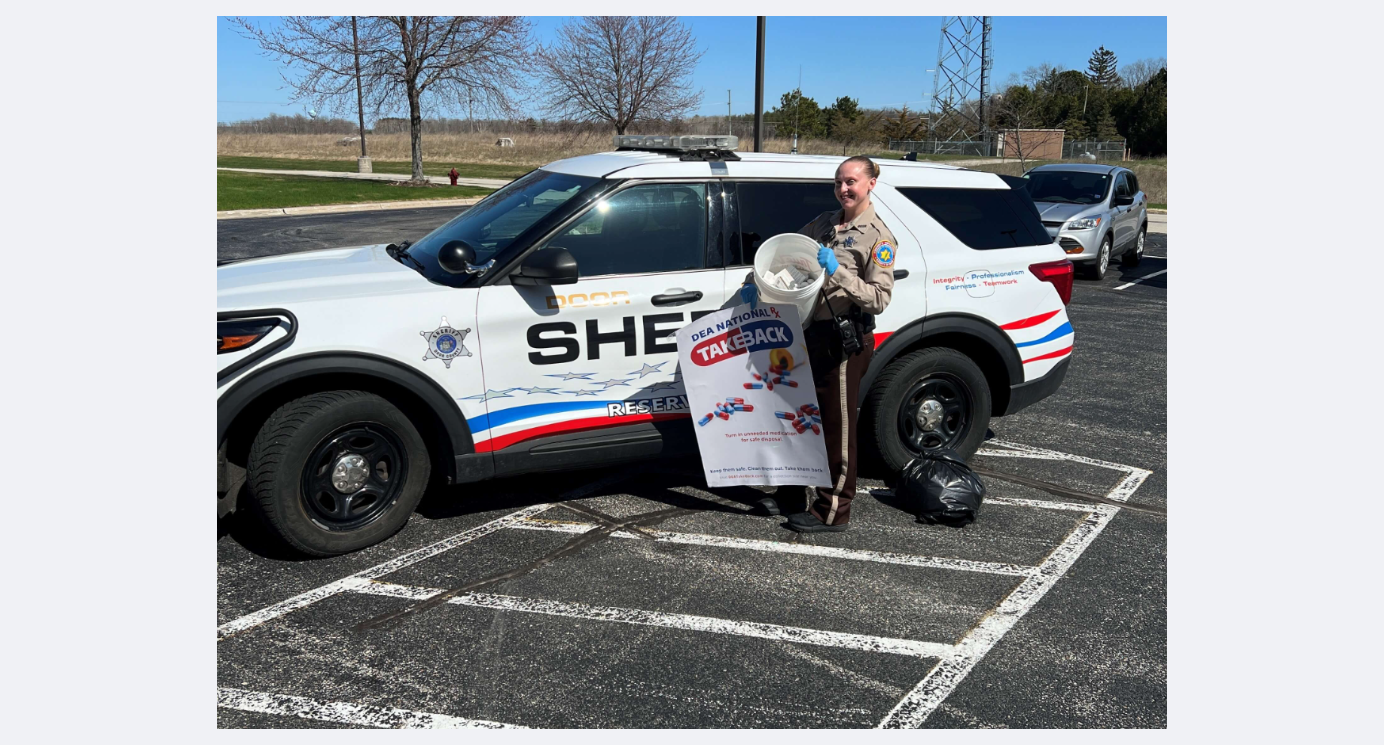 Delilah Rose standing in front of a reserve deputy squad car, holding a National Drug Take Back Day poster alongside an evidence bin filled with collected prescription drugs.