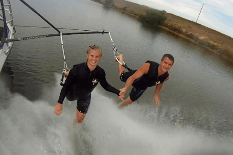 Two individuals water skiing, holding onto a tow bar connected to a boat, with a calm waterway and greenery in the background.