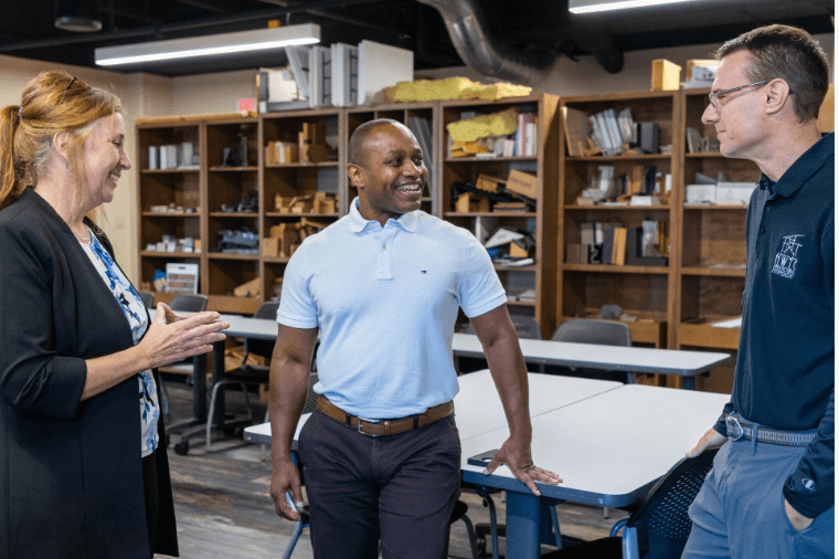 Three adults conversing joyfully in a classroom setting with shelves and tables visible in the background.