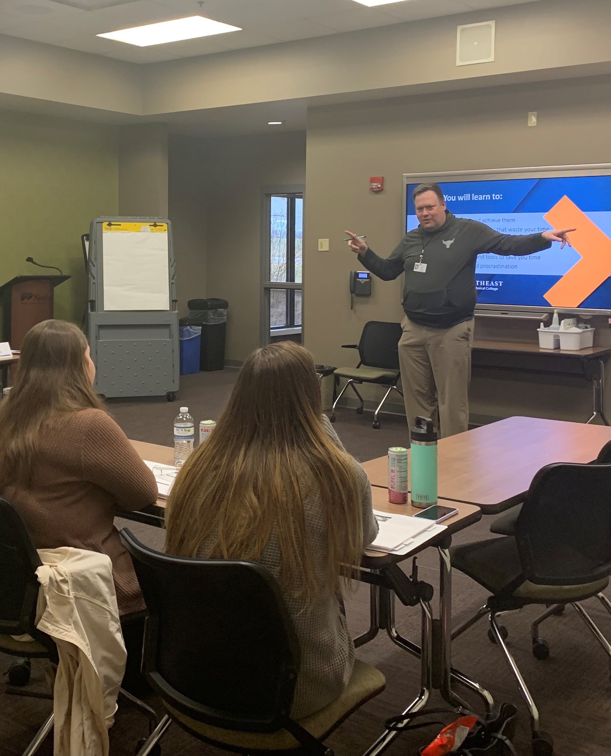 A group of people sitting at tables in a classroom watching a speaker present on a screen. 