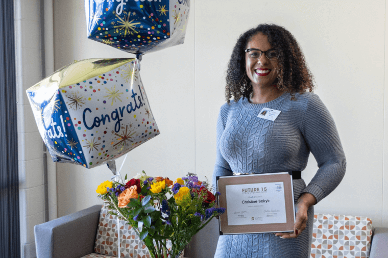 A woman in a gray dress stands next to balloons that say, “Congrats!” and a bouquet of flowers while holding a certificate that says, “Future 15.”