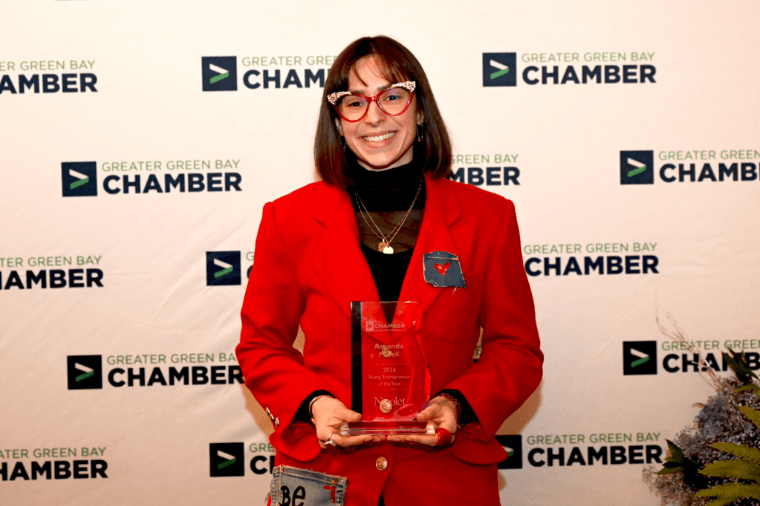 A person wearing a red blazer holds an award in front of a backdrop with the "Greater Green Bay Chamber" logo.