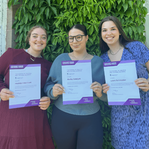 Three people smiling and holding certificates in front of green foliage.