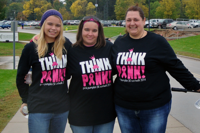 Three people posing together outdoors, all wearing black t-shirts with "Think Pink" in large pink letters for a breast cancer awareness event. There's a park and parking area in the background.