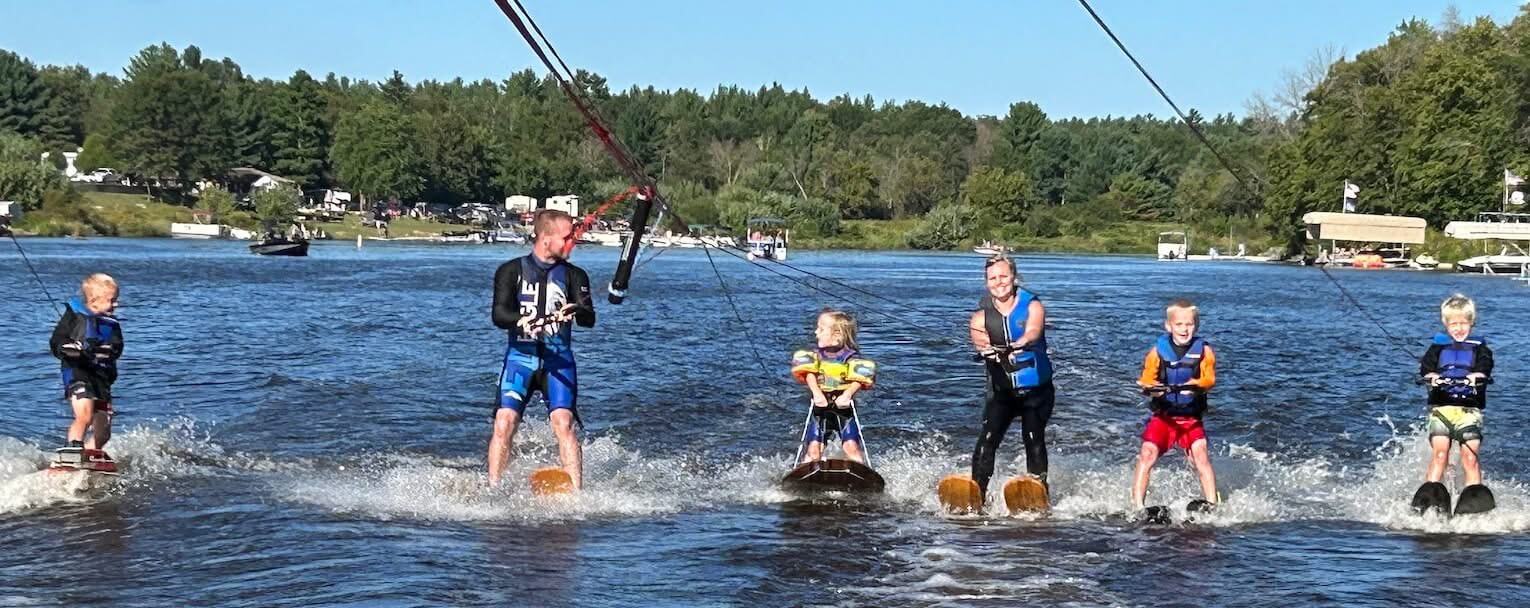 Five individuals water skiing on a sunny day, each holding onto cables and wearing life jackets. Water and trees visible in the background.