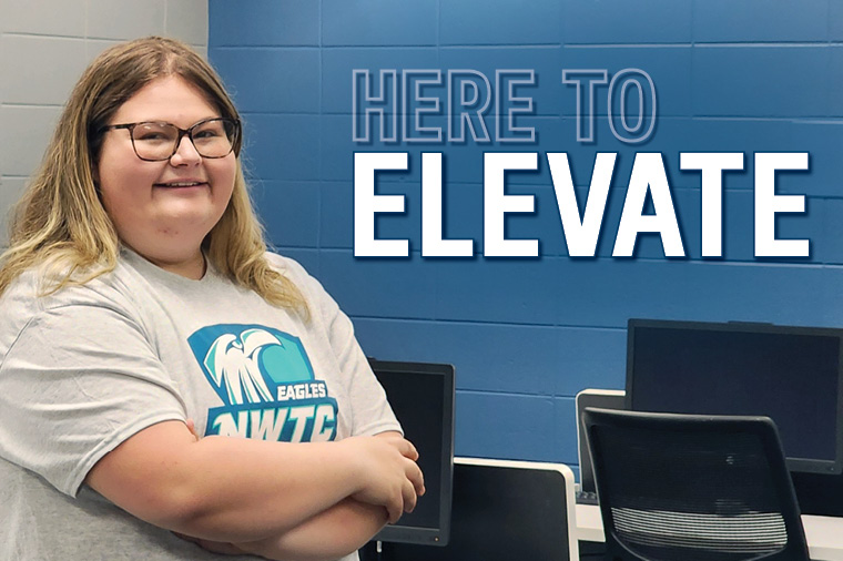 Student stands in front of computers with Here to Elevate text