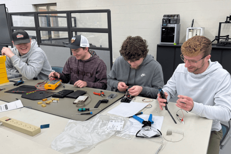 Four students are engaged in assembling electronic components at a workshop table, smiling and focused on their tasks.