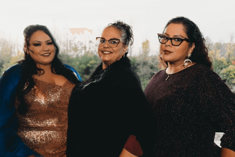 Three women posing together for a photo, each dressed in elegant attire, with a blurred natural background.