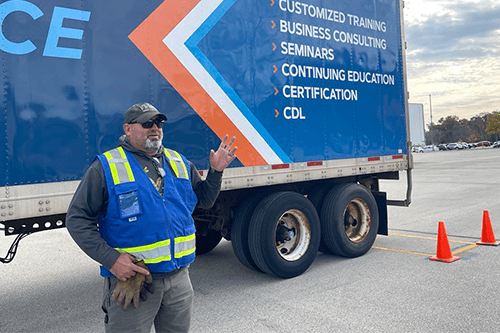 Person wearing a high‑visibility safety vest stands beside a branded semi‑truck trailer listing training services, including customized training, business consulting, seminars, continuing education, certification, and CDL.